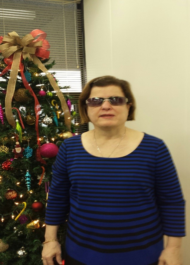 Woman in a blue shirt and glasses smiling and standing next to a Christmas tree.